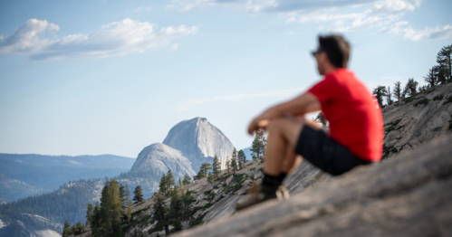 On a sheer rock face, a hiker in black shorts and a red t-shirt sits and looks out at Yosemite's Half Dome in the distance.