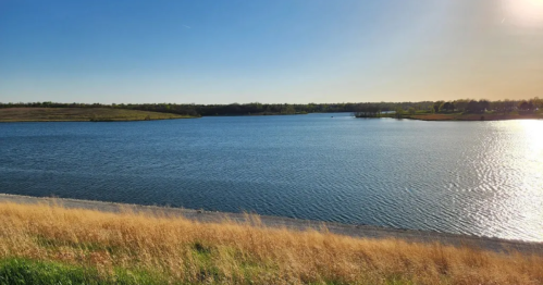 A serene lake surrounded by green hills and golden grass under a clear blue sky. Sunlight reflects on the water.