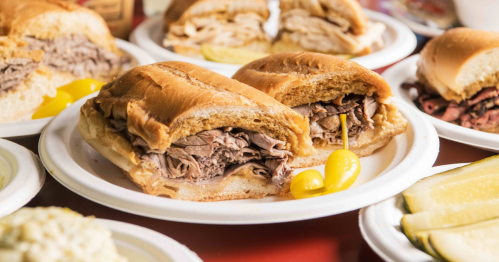 Plates of delicious food crowd a red table, arranged around a beef french dip sandwich.