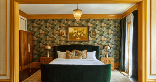 A stylish bedroom featuring a green velvet bed, patterned wallpaper, and elegant decor with a chandelier and artwork.