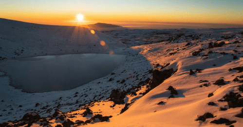 In a snowy landscape, a small alpine lake stands out at sunset.