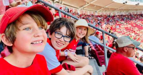 Three children in red shirts smile while sitting in a stadium filled with fans, enjoying a lively event.
