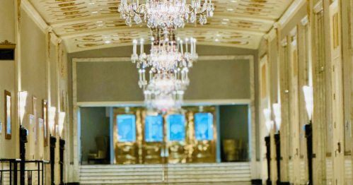 Elegant hallway with ornate chandeliers and golden doors at the end, featuring marble walls and soft lighting.