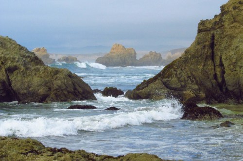 Waves crash against rocky shores, with misty cliffs and distant formations under a cloudy sky.