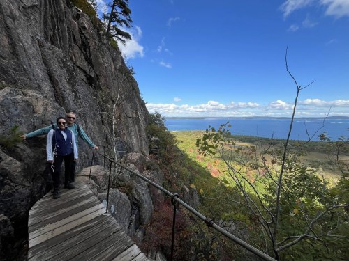 Two people stand on a wooden path along a rocky cliff, overlooking a scenic landscape with trees and a body of water.
