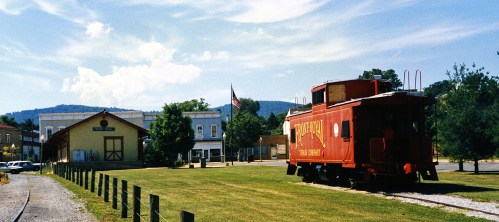 A red caboose sits on grass near a historic train depot, with mountains and an American flag in the background.