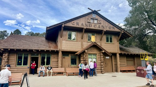 Log cabin-style building with "Grand Canyon" sign, people gathered outside, and benches in front under a blue sky.