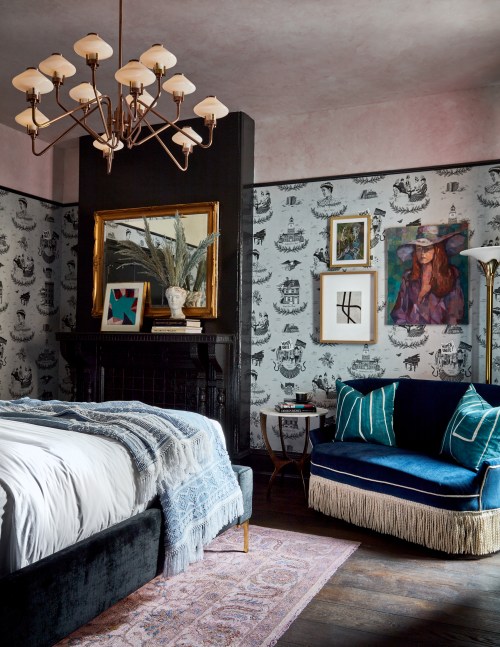 A stylish bedroom featuring a bed, a blue fringed sofa, artwork, and a chandelier against patterned wallpaper.