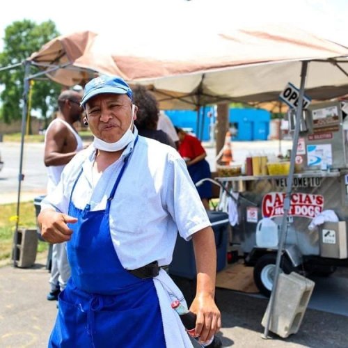 A vendor in a blue apron and cap stands outside a food cart, smiling in a sunny outdoor setting.