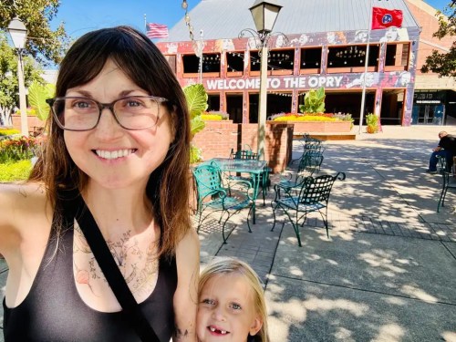 A smiling woman and a child pose outdoors in front of the Opry, with greenery and seating visible around them.