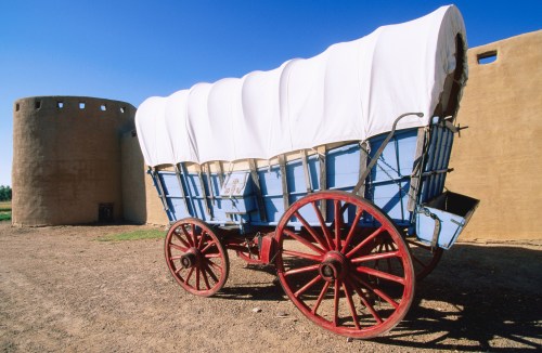 A blue covered wagon with red wheels parked near a clay structure under a clear blue sky.