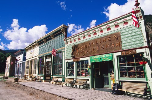 Historic storefronts line a wooden boardwalk under a blue sky with fluffy clouds, featuring colorful signs and American flags.