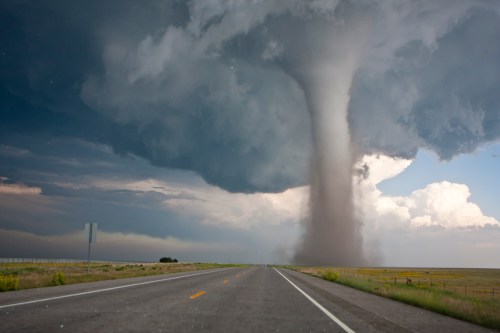 A powerful tornado descends from dark storm clouds over an empty road in a rural landscape.