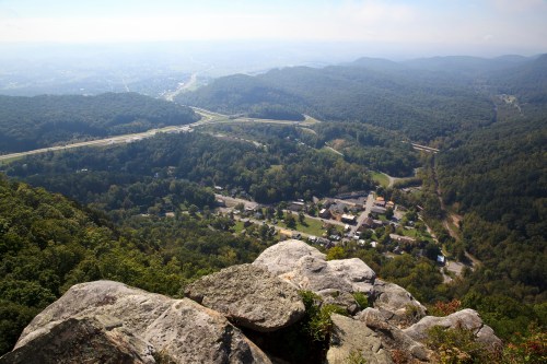 A panoramic view from a rocky overlook, showcasing a valley with a small town and winding roads surrounded by green hills.