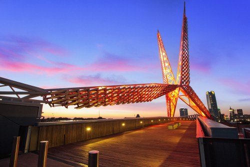 A striking bridge with a unique design, illuminated in orange against a colorful sunset sky. City skyline in the background.