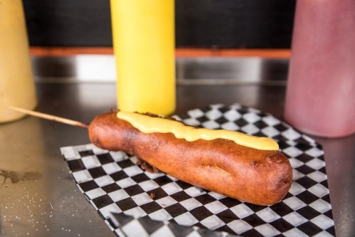 A golden-brown corn dog drizzled with yellow mustard on a black and white checkered paper.