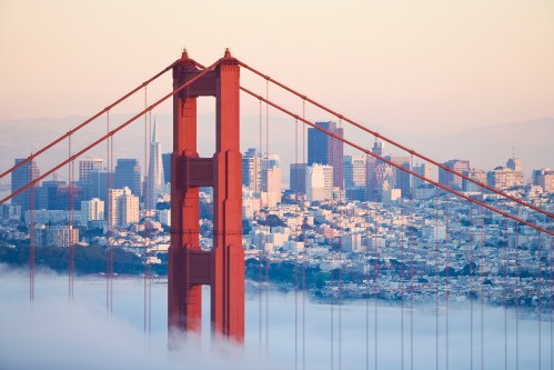 Golden Gate Bridge towers over a foggy San Francisco skyline at sunrise, with city buildings in the background.