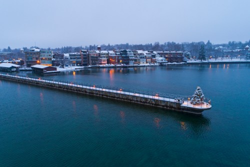 A snowy waterfront scene with a pier leading to a quaint town, illuminated buildings, and a calm, icy lake.