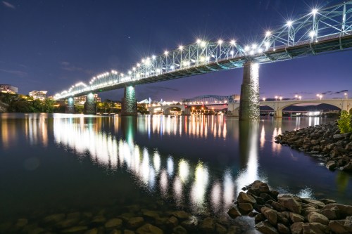 A brightly lit bridge spans a calm river at night, reflecting lights on the water's surface.