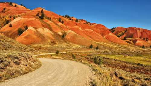 A winding dirt road leads through vibrant red and orange hills under a clear blue sky. Green fields are visible below.