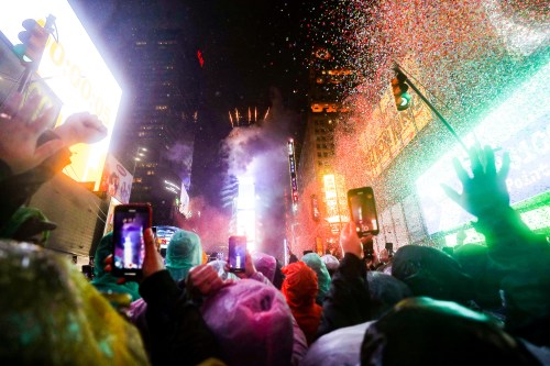Crowd in rain ponchos celebrating New Year's Eve in Times Square, with confetti and bright lights illuminating the night.