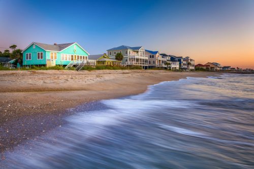 Colorful beach houses line a sandy shore, with gentle waves lapping at the beach under a twilight sky.