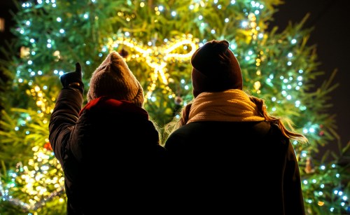 Two people in winter attire admire a brightly lit Christmas tree adorned with lights and a large bow.