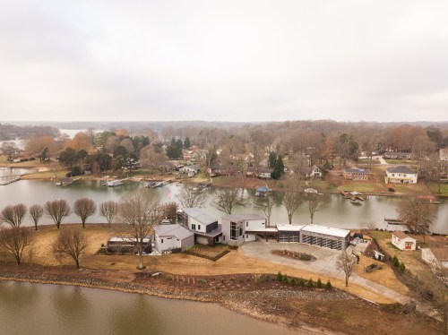 Aerial view of a modern house by a lake, surrounded by trees and residential areas, with boats along the shore.