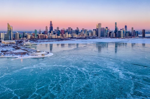 Aerial view of a frozen lake with a city skyline at sunset, featuring skyscrapers and a pastel sky.