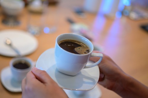 A person holding a white coffee cup on a saucer, with a dark beverage inside, against a blurred background.