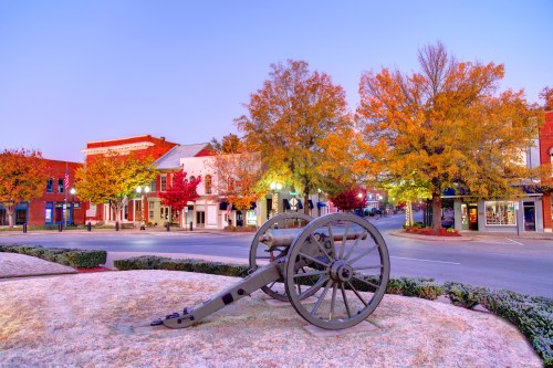 A historic cannon sits on a grassy area, surrounded by colorful autumn trees and charming buildings at dusk.