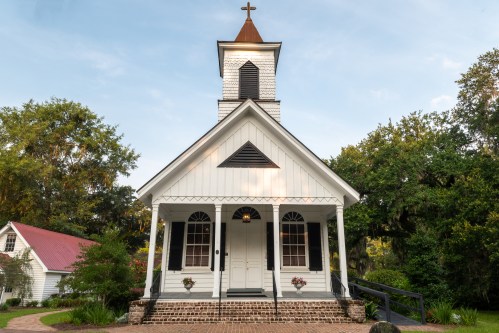A charming white church with a steeple, surrounded by greenery and a brick pathway, under a clear blue sky.