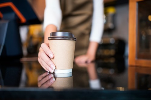 A barista holds a disposable coffee cup at a café counter, with a blurred background of the shop.