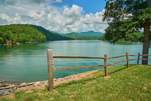 A serene lake surrounded by green hills and trees, with a wooden fence in the foreground under a partly cloudy sky.