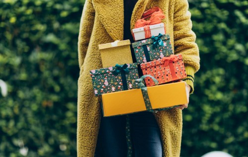 A person in a yellow coat holds a stack of colorful wrapped gifts against a green background.