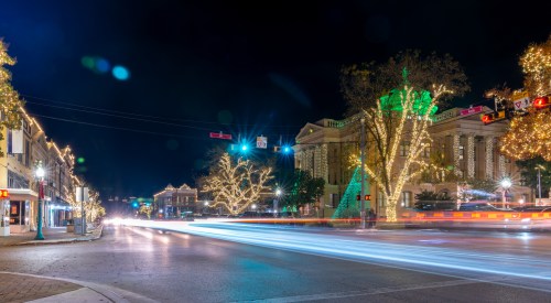 A festive street scene at night, adorned with colorful lights and decorated trees, showcasing holiday spirit.