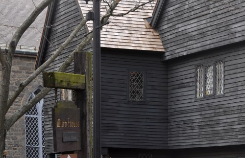 A dark wooden house with a steep roof and diamond-patterned windows, next to a sign reading "Witch House."