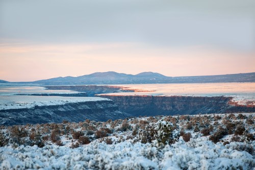 A snowy landscape with a canyon, distant mountains, and a soft pastel sky at dawn.