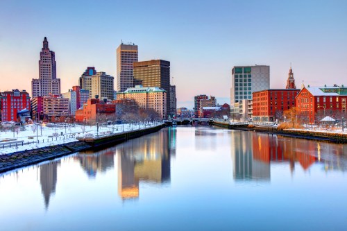 City skyline reflecting on a river at dawn, featuring modern and historic buildings with a snowy landscape.