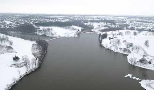 Aerial view of a snowy landscape with a river, trees, and a distant residential area under a gray sky.
