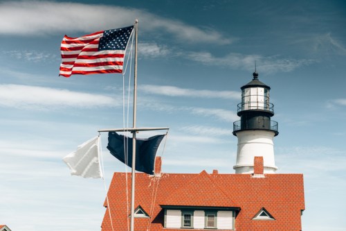 A lighthouse with a black lantern stands beside an American flag and two other flags against a blue sky.