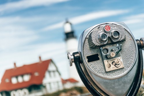 A coin-operated viewing telescope in the foreground, with a lighthouse and house blurred in the background.