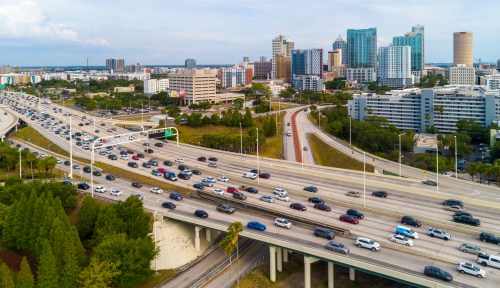 Aerial view of a busy highway with traffic, surrounded by urban buildings and greenery in a cityscape.