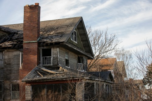 An abandoned, dilapidated house with a crumbling roof and overgrown vegetation, under a cloudy sky.