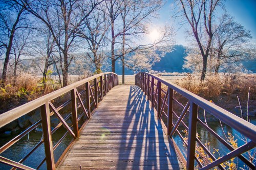 A wooden bridge spans a serene river, surrounded by frosty trees and a bright sun shining in a clear blue sky.