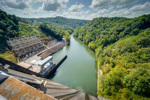 Aerial view of a dam on a river, surrounded by lush green hills and a partly cloudy sky.