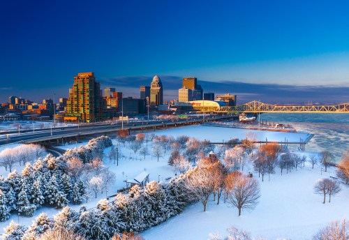 A winter cityscape of Louisville, Kentucky, featuring snow-covered trees and buildings under a clear blue sky.