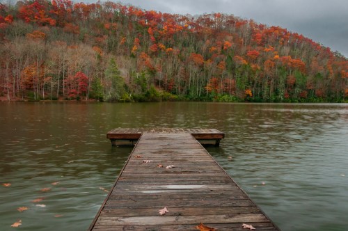 A wooden dock extends into a calm lake, surrounded by trees with vibrant autumn foliage.