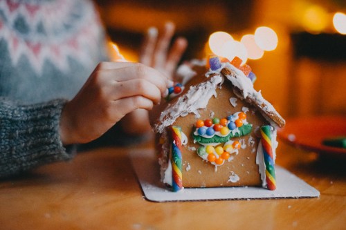 A person decorates a gingerbread house with colorful candies and icing, with warm lights in the background.