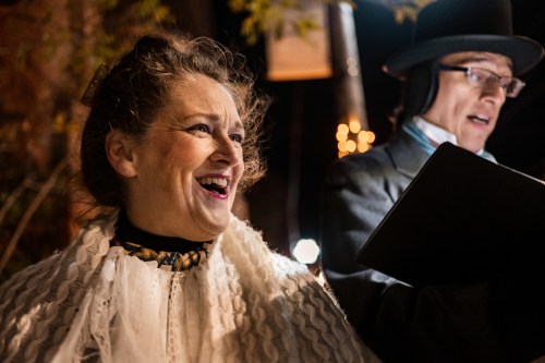 A woman in historical attire smiles brightly while a man in a top hat reads from a book nearby, set at night.